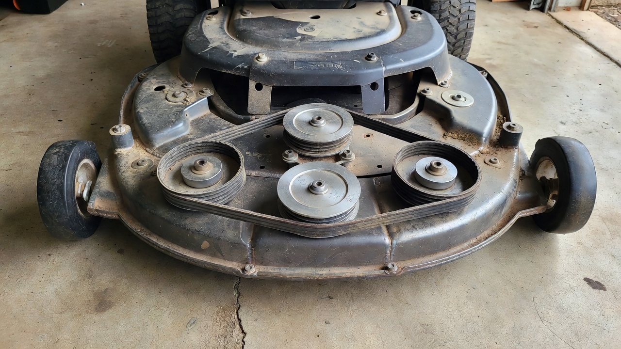 Underside of a riding lawn mower deck showing multiple metal pulleys and rubber drive belt routing