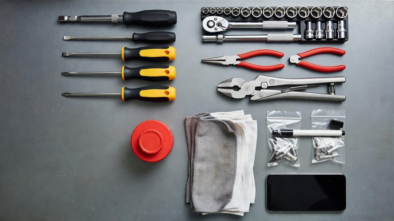 Overhead flat-lay of small engine repair tools including ratchet, nut drivers, needle-nose pliers, fuel container, shop rags, and zip-lock bags on a gray workbench