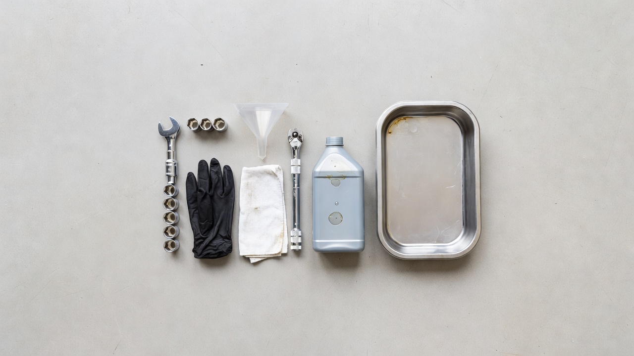 Tools needed for a lawn mower oil change laid out on a workshop floor including wrench, gloves, funnel, and drain pan