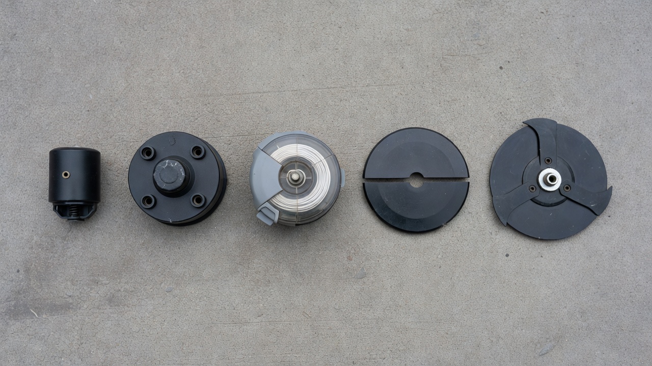 Five different strimmer head types arranged side by side on a concrete surface showing bump-feed, auto-feed, fixed-line and blade designs