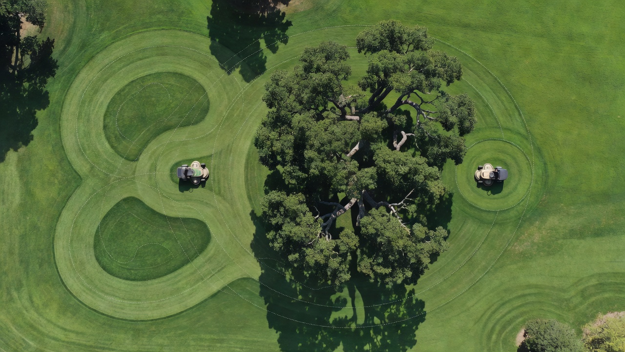 Aerial comparison of wide-turn conventional mower versus zero-turn mower path around trees on a commercial lawn